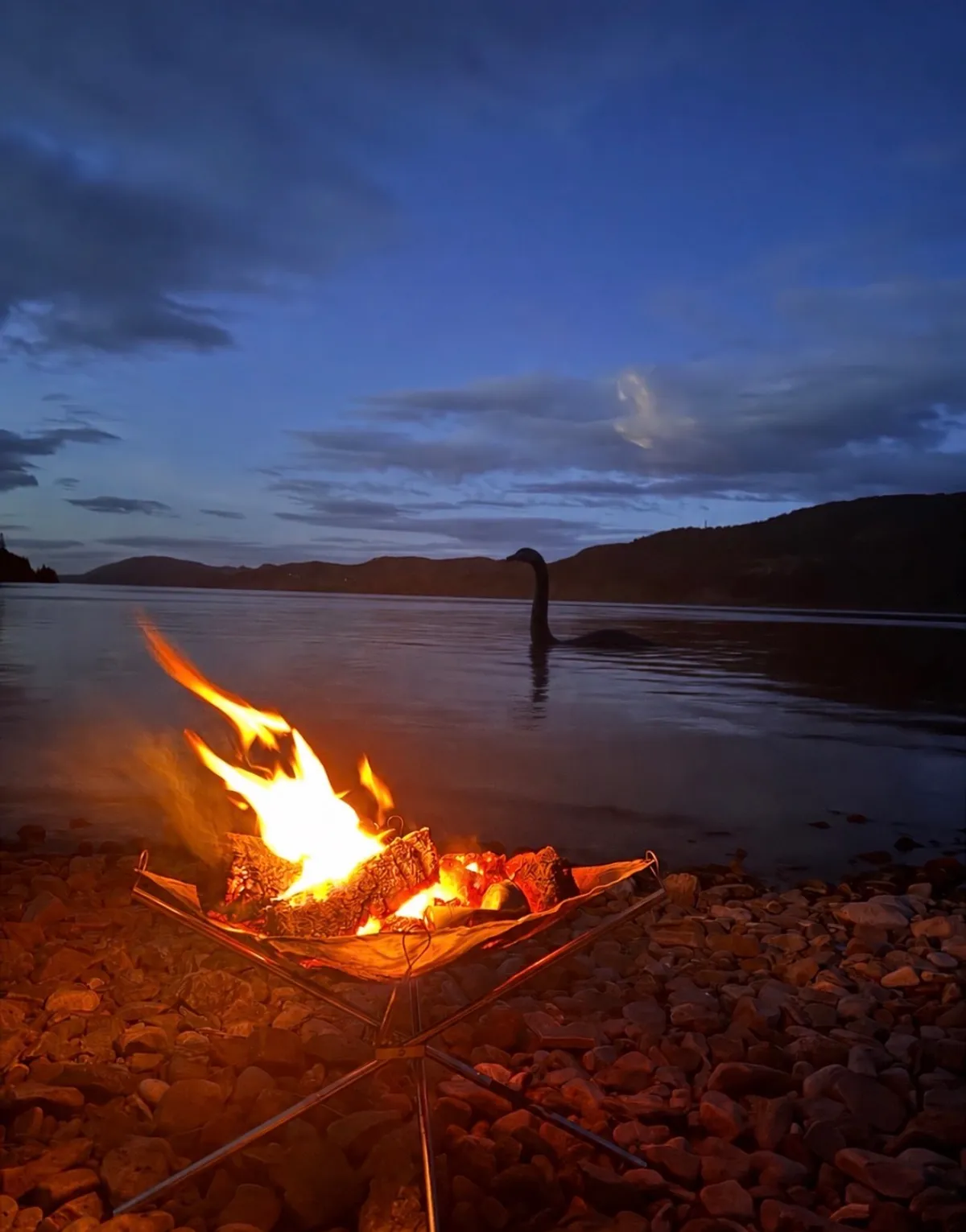 A campfire on the shore of Loch Ness at dusk with a Nessie silhouette in the water